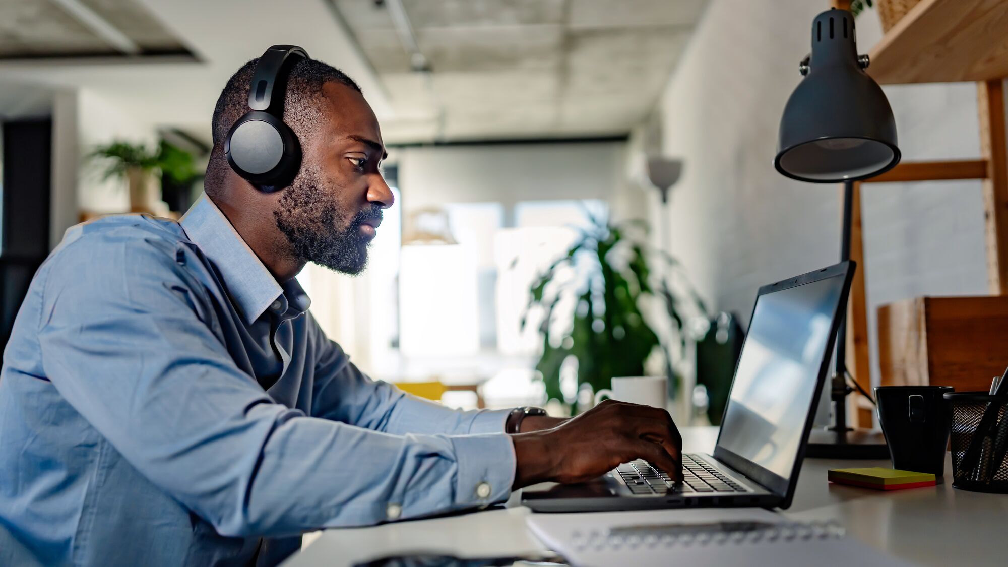 Focused man wearing headphones, working on his laptop in a modern, well-lit home office environment.